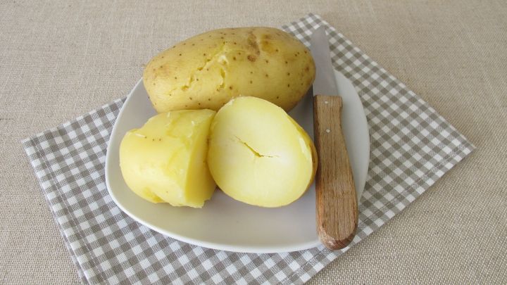 Ein Teller mit Pellkartoffeln, einige geschnitten, liegt auf einer karierten Serviette, w&auml;hrend ein Holzmesser daneben liegt.