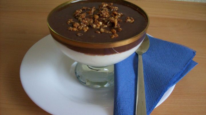 Ein Dessertglas mit einer Schicht Frischk&auml;se-Creme, bedeckt mit Schokolade und gehackten N&uuml;ssen, steht auf einem Teller.