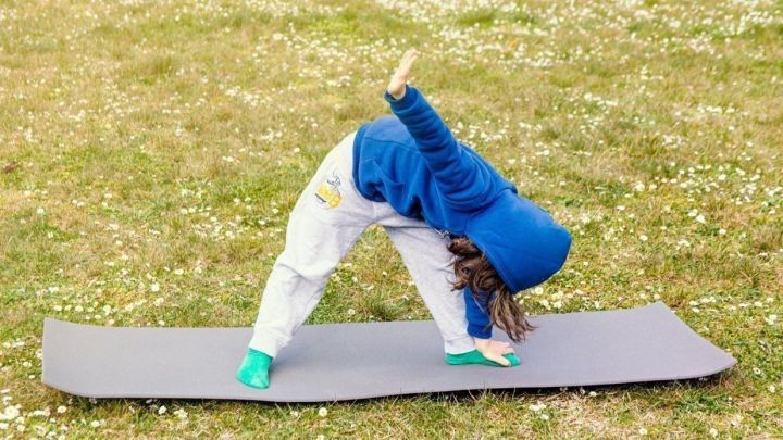 Ein Kind macht mit einem blauen Hoodie und grauen Jogginghosen auf einer Matte eine Yoga-&Uuml;bung in einer bl&uuml;henden Wiese.
