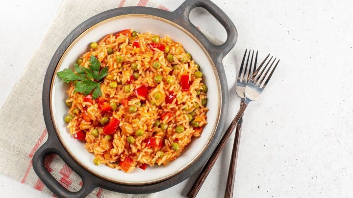 Eine Sch&uuml;ssel Djuvec Reis ist gef&uuml;llt mit buntem Gem&uuml;se, auf einem Tisch serviert mit Besteck und einem dekorativen Tuch.