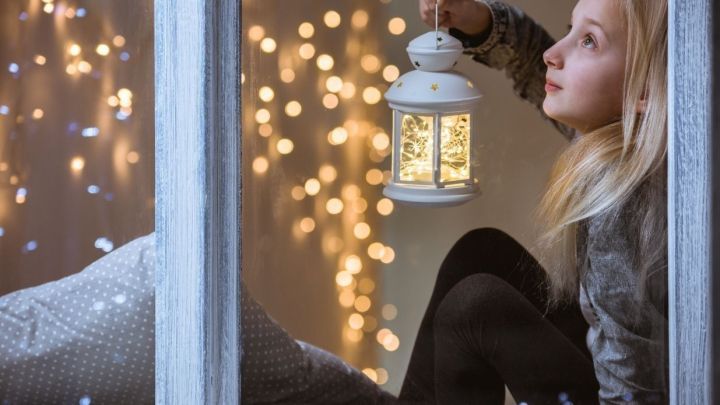 Ein M&auml;dchen sitzt am Fenster und h&auml;lt sp&auml;terns eine leuchtende Laterne, w&auml;hrend im Hintergrund warme Lichter funkeln.
