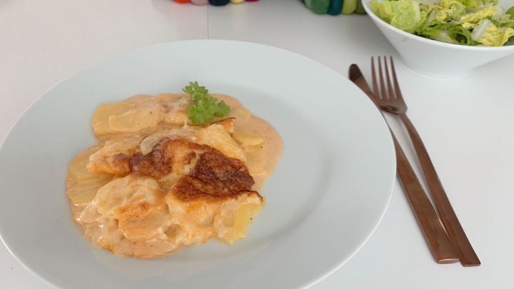 Ein cremiges Kartoffelgratin liegt auf einem wei&szlig;en Teller, umgeben von frischem Salat in einer Sch&uuml;ssel auf dem Tisch.