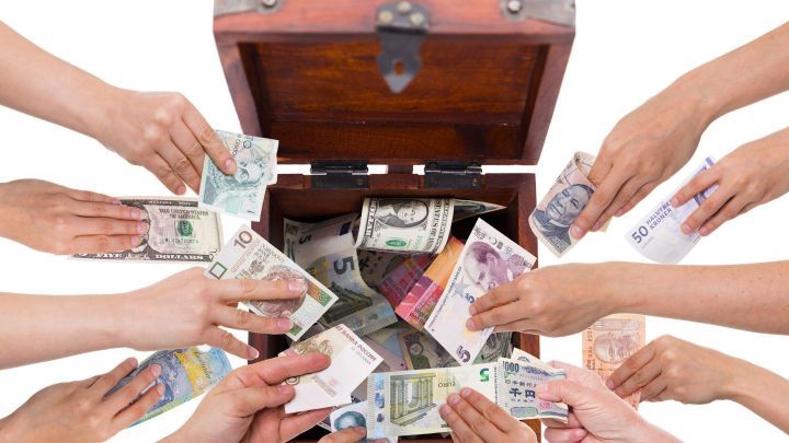 Eine Gruppe von H&auml;nden legt Geldscheine in eine offene Schatzkiste, die bereits mit verschiedenen W&auml;hrungen gef&uuml;llt ist.