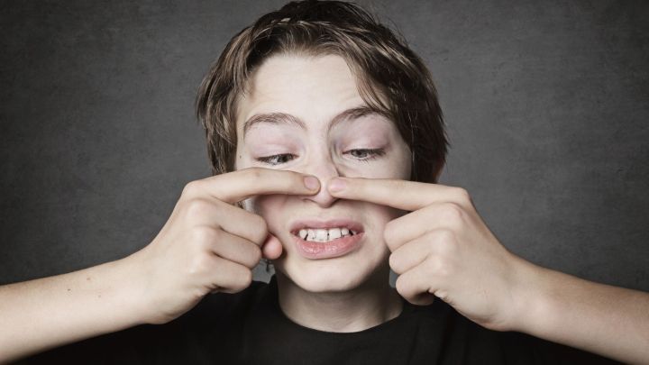 Ein Junge dr&uuml;ckt mit seinen Fingern auf seine Nase, wahrscheinlich um Pickel auszudr&uuml;cken, vor einem grauen Hintergrund.