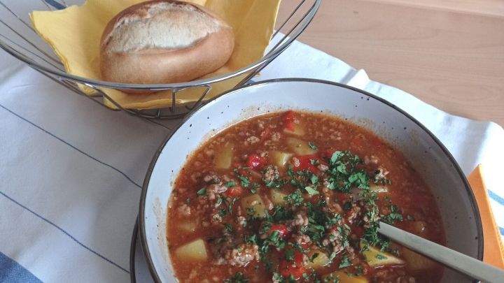 Ein Puszta-Topf mit Hackfleisch ist in einer Sch&uuml;ssel angerichtet, serviert mit einem Br&ouml;tchen im Korb, auf einem Tisch.