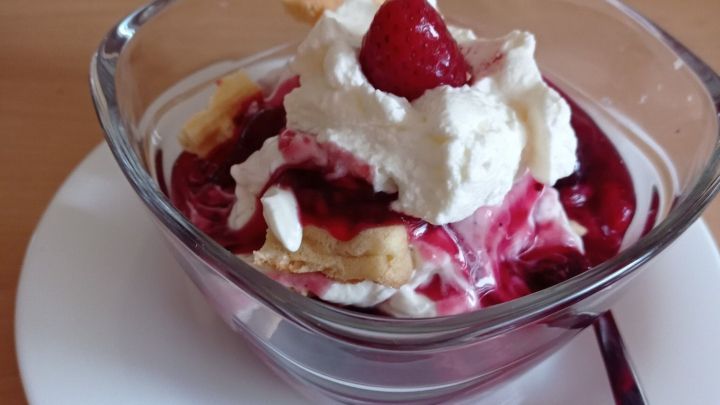 Eine Sch&uuml;ssel mit Quarkspeise wird mit roter Gr&uuml;tze, Sahne und einer Waffel dekoriert, auf einem Holzuntergrund pr&auml;sentiert.