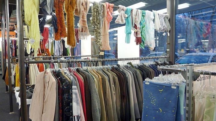 Bunte Kleidung und Schals h&auml;ngen ordentlich in einem Sozialkaufhaus, w&auml;hrend durch gro&szlig;e Fenster Tageslicht str&ouml;mt.