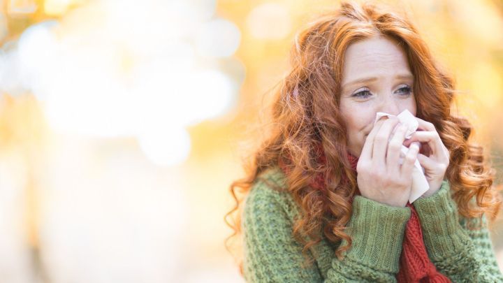 Eine Frau mit roten Locken h&auml;lt ein Taschentuch, w&auml;hrend sie in einem herbstlichen Park leidet unter Heuschnupfen-Symptomen.