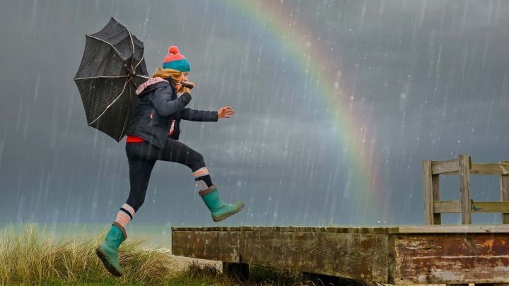 Eine Person springt l&auml;chelnd mit einem Regenschirm &uuml;ber eine Holzplanke, w&auml;hrend es regnet und ein Regenbogen zu sehen ist.
