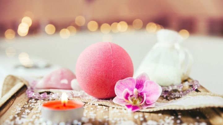 Eine rosa Badebombe liegt auf einem Holztablett, umgeben von einem Teelicht, Blumen und dekorativem Salz in einer entspannenden Umgebung.