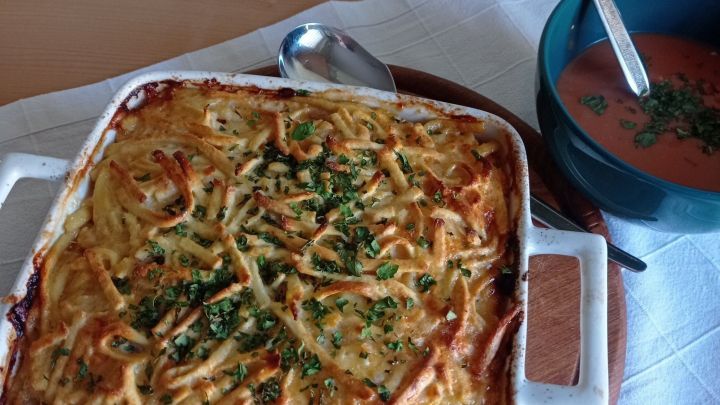 Ein goldbrauner Nudel-Putenfleisch-Auflauf liegt auf einem Holzbrett, umgeben von einer Sch&uuml;ssel mit Tomatensuppe.