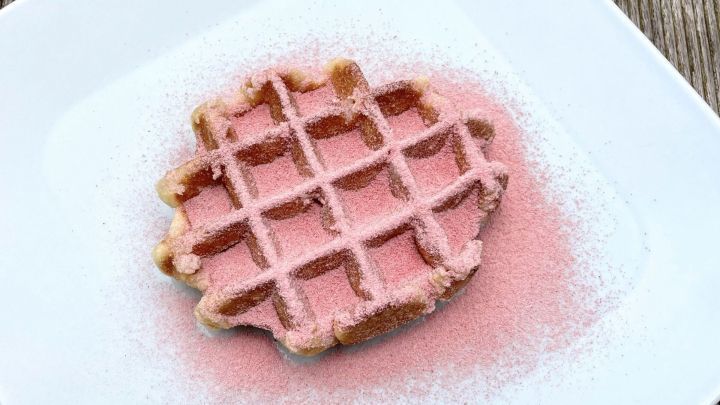 Eine Waffel liegt auf einem Teller, bedeckt mit rosa Erdbeerpuderzucker, vor einer Holztischoberfl&auml;che.