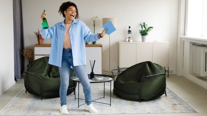 Eine fr&ouml;hliche Person h&auml;lt eine Flasche Reinigungsmittel und ein blaues Buch in einem hellen Raum mit Pflanzen und M&ouml;beln.