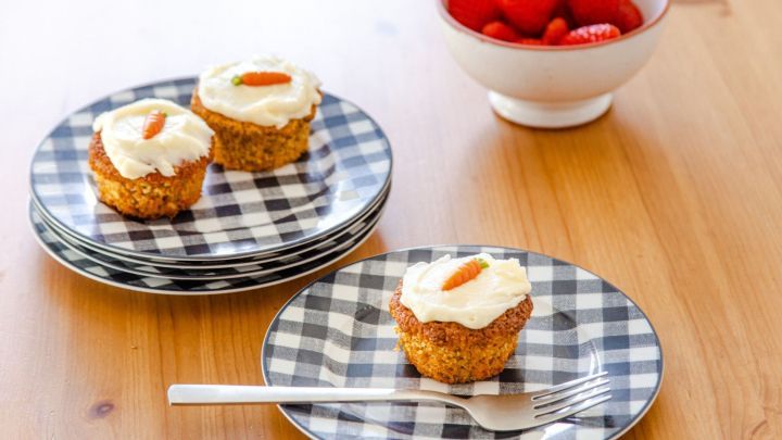 Saftige Karottenmuffins stehen auf karierten Tellern, umgeben von einem Obstsch&uuml;ssel mit frischen Erdbeeren auf Holztisch.