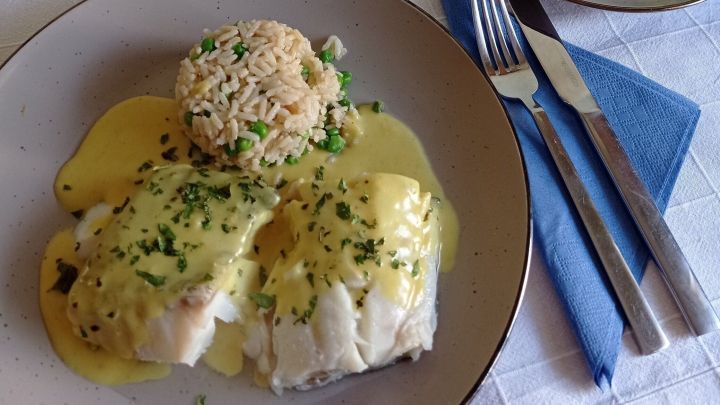 Kabeljau in Curryso&szlig;e liegt auf einem Teller mit Reis und Gem&uuml;se, umgeben von Besteck und einer Tischdecke.