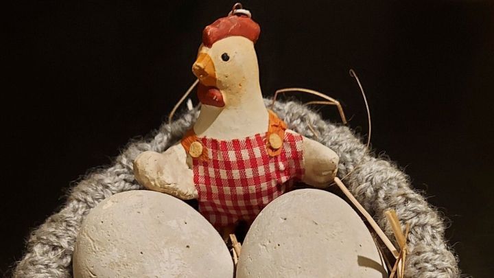 Eine dekorative Henne sitzt in einem offenen, gestrickten Nest und bewacht drei beige Eier auf Stroh.