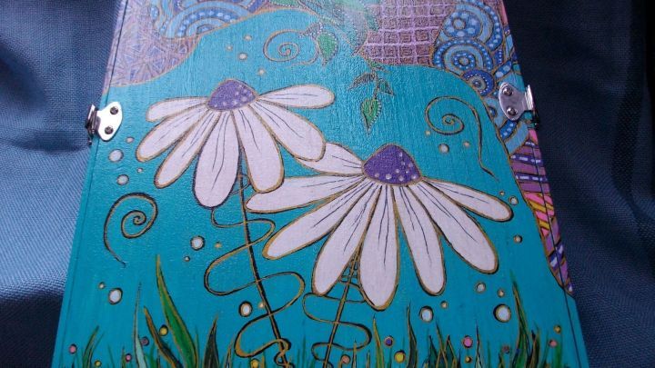 Ein bemalter Besteckkasten mit bunten Blumen und spiralf&ouml;rmigen Details steht auf einem blauen Hintergrund.