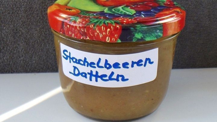 Ein Glas mit Stachelbeer-Dattel Marmelade steht auf einem Tisch, umgeben von einer schlichten grauen Wand.
