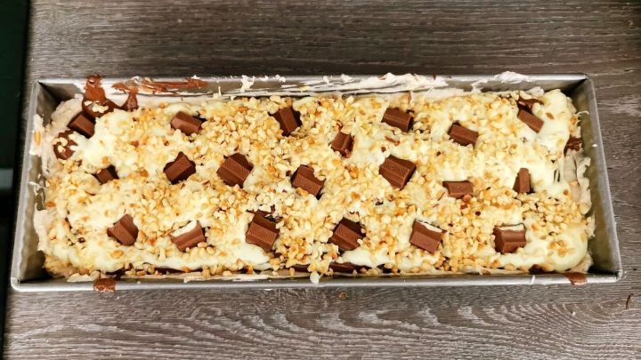 Ein &uuml;ppiger Kinderriegel-Kuchen liegt in einer Backform, bedeckt mit Schokoladenst&uuml;cken und gehackten N&uuml;ssen.