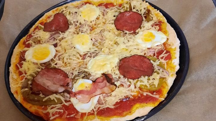 Eine Pizza mit Ei liegt auf einem Backblech, goldbraun gebacken, belegt mit K&auml;se, Speck und mehreren Eiern.