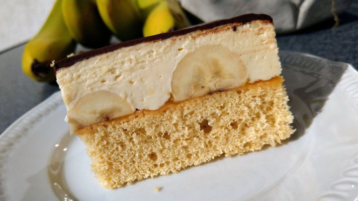 Ein St&uuml;ck Bananenschnitten mit Bananenscheiben und einer cremigen Schicht liegt auf einem wei&szlig;en Teller vor einigen Bananen.