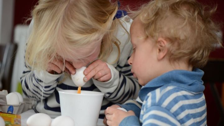 Ein Kind bl&auml;st ein Ei &uuml;ber einer wei&szlig;en Sch&uuml;ssel aus, w&auml;hrend ein anderes Kind neugierig zuschaut. Eier werden vorbereitet.