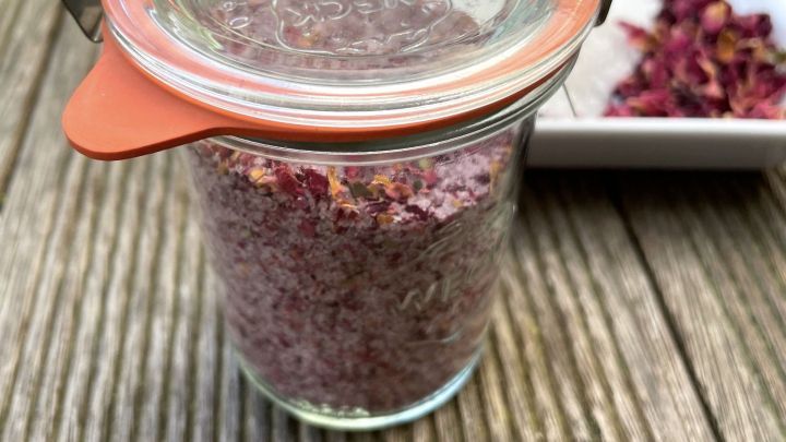 Ein Glas mit Rosensalz steht auf einem Holztisch, w&auml;hrend getrocknete Rosenbl&auml;tter daneben liegen.