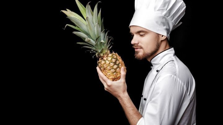 Ein Koch h&auml;lt eine frische Ananas in der Hand, w&auml;hrend er nachdenklich auf die Frucht blickt, vor einem schwarzen Hintergrund.
