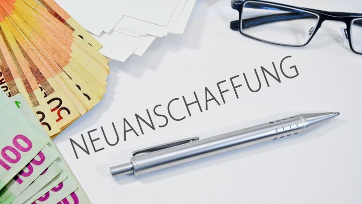 Ein Stapel Geldscheine liegt neben einem Stift auf einem Tisch mit dem Wort &bdquo;NEUANSCHAFFUNG&ldquo;, um Kaufentscheidungen zu treffen.