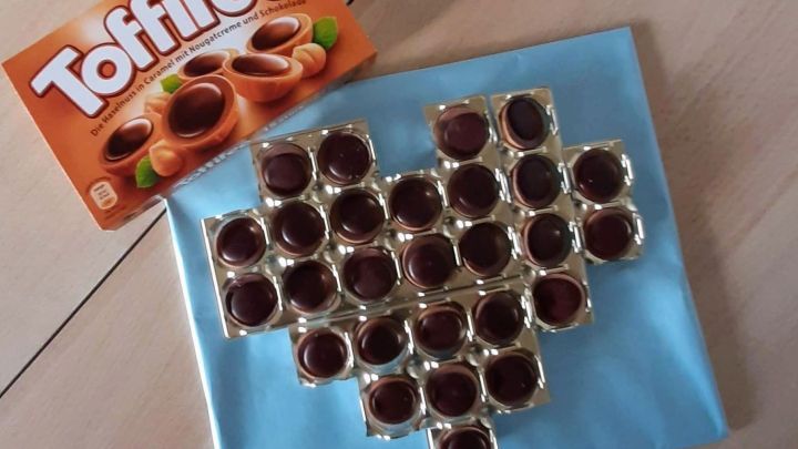 Auf einem blauen Untergrund sind Toffifee-Karamellbonbons in Herzform angeordnet, w&auml;hrend eine Verpackung daneben liegt.