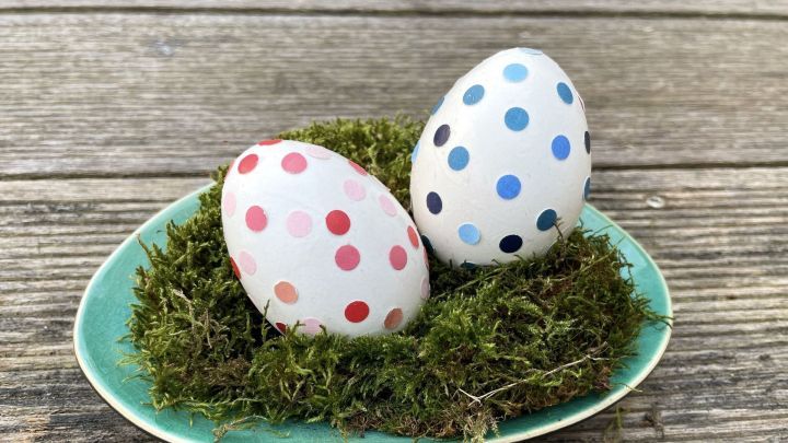 Auf einer t&uuml;rkisfarbenen Platte liegen zwei Ostereier mit bunten Punkten auf gr&uuml;nem Moos, umgeben von nat&uuml;rlichem Holz.
