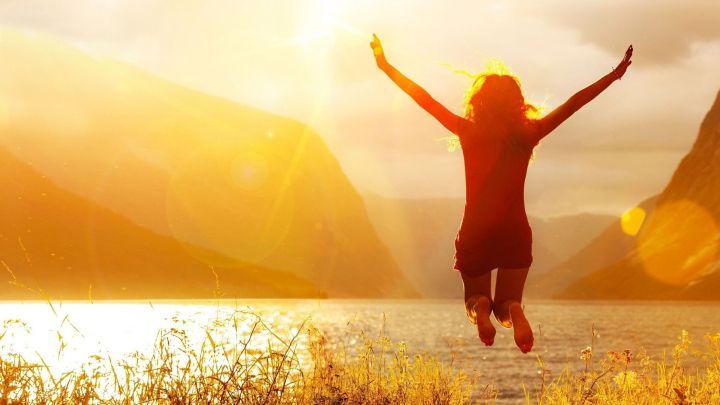 Eine Person springt freudig in die Luft, umgeben von einer idyllischen, sonnigen Landschaft mit Bergen und Wasser.