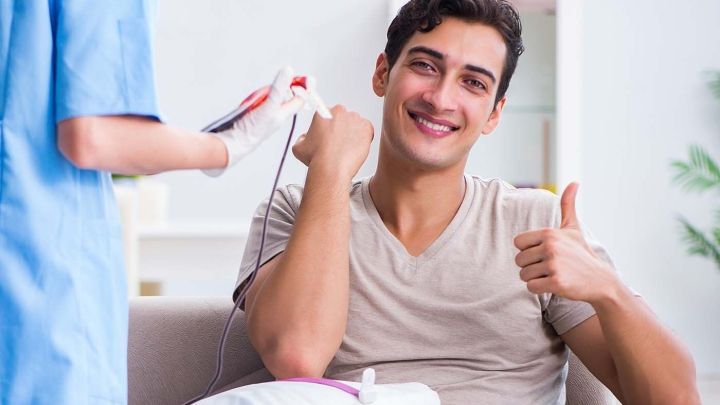 Ein junger Mann sitzt auf einem Sofa und spendet Stammzellen. Er l&auml;chelt und zeigt Daumen nach oben, w&auml;hrend eine Fachkraft ihm dabei hilft.