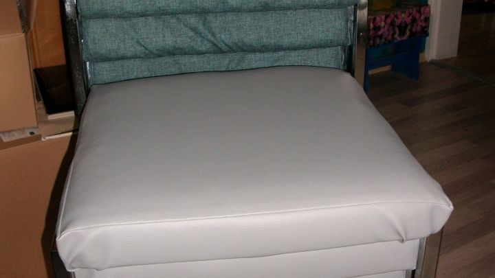 Ein farbenfroher Stuhl mit grauem Bezug steht in einem Raum, umgeben von Regalen und Kisten, zum Neubeziehen bereit.