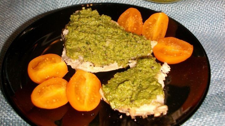 Auf einem schwarzen Teller liegen zwei Brote mit Walnuss-B&auml;rlauch-Pesto, umgeben von gelben Tomatenscheiben. Jar mit Pesto im Hintergrund.