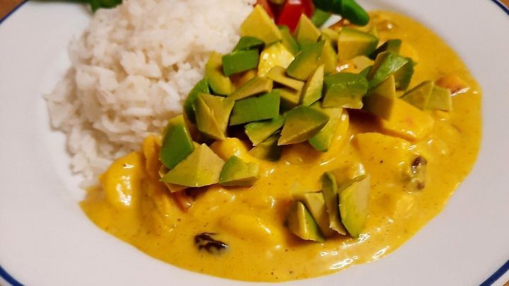 Saftiges H&auml;hnchencurry mit Avocado liegt neben Reis und frischem Salat auf einem Teller aus Holz.