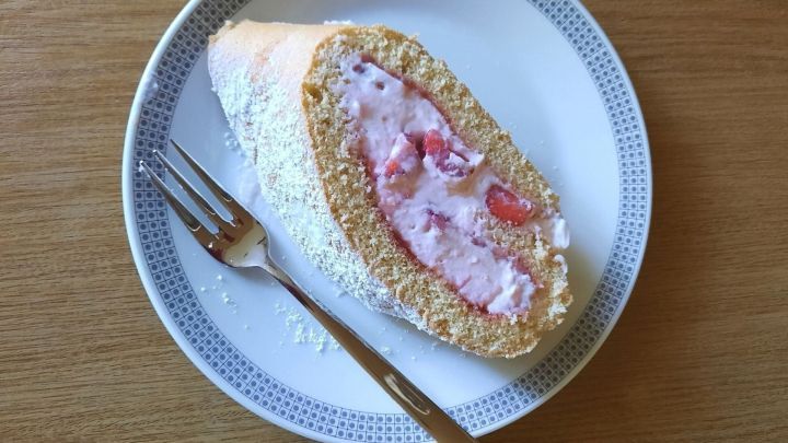 Eine Biskuitrolle mit Erdbeeren liegt auf einem Teller, dekoriert mit Puderzucker, w&auml;hrend eine Gabel daneben liegt.