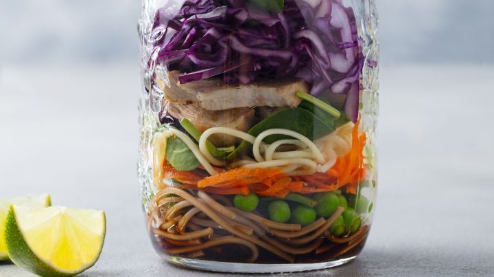 Ein Glas mit buntem Gem&uuml;se, Nudeln und Sauce wird in einer K&uuml;chenumgebung pr&auml;sentiert, daneben eine Limette.