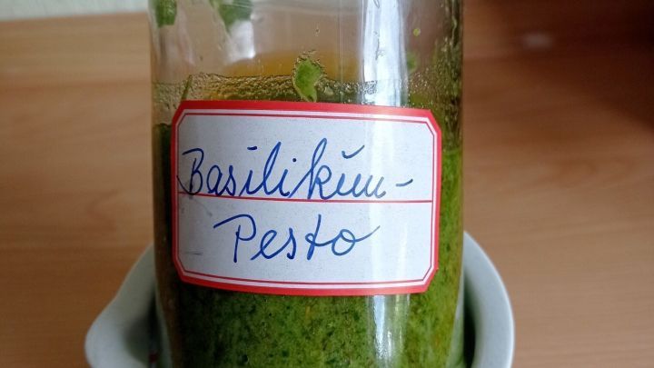 Ein Glas Basilikum-Pesto steht in einer wei&szlig;en Schale auf einer Holzoberfl&auml;che und zeigt eine leuchtend gr&uuml;ne Farbe.