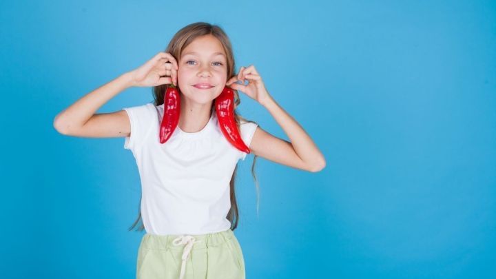 Ein M&auml;dchen in einem wei&szlig;en T-Shirt und hellgr&uuml;nen Shorts h&auml;lt rote Paprika neben ihrem Gesicht und l&auml;chelt vor einem blauen Hintergrund.