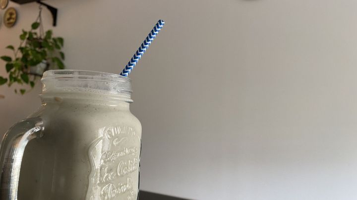 Ein Bananenshake in einem Glas mit einem gestreiften Strohhalm steht auf einem Tisch in einem hellen Raum mit Pflanzen.