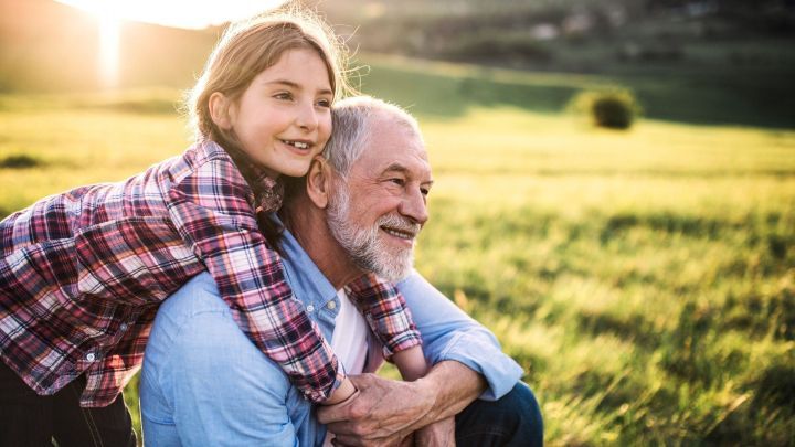 Ein Kind umarmt seinen Gro&szlig;vater von hinten, beide l&auml;cheln gl&uuml;cklich in einer sonnigen Wiese.