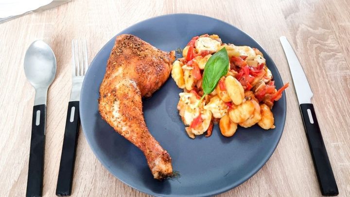 Ein gebratenes H&auml;hnchenschenkel liegt neben einem bunten Gnocchi-Salat auf einem blauen Teller, umgeben von Besteck.
