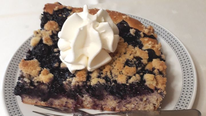 Ein St&uuml;ck Streuselkuchen mit Heidelbeeren und Sahnehaube liegt auf einem Teller, dekoriert auf einem wei&szlig;en Tisch.