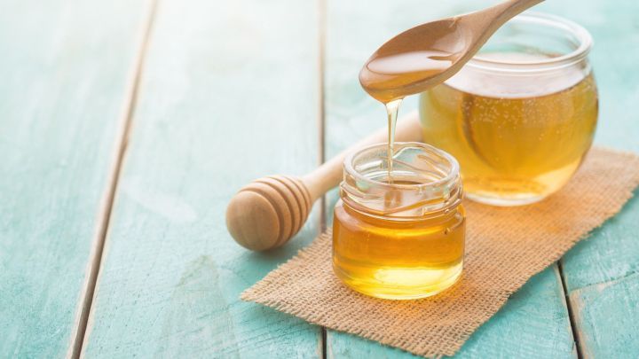 Ein L&ouml;ffel Honig wird &uuml;ber ein kleines Glas mit Honig gehalten, auf einer Holzplatte und einem Jutesack im Hintergrund.