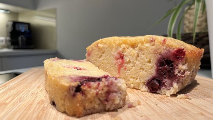 Ein Grie&szlig;kuchen mit Beeren liegt auf einem Holzbrett, w&auml;hrend ein St&uuml;ck davon bereits abgeschnitten ist.