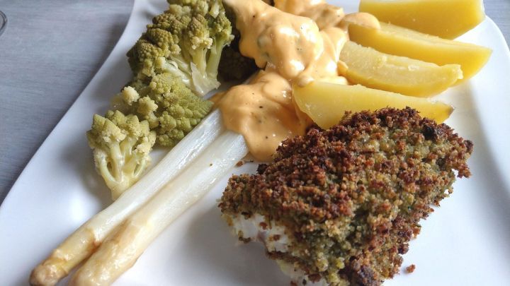 Kabeljaufilet liegt knusprig gebacken auf einem Teller, umgeben von Gem&uuml;se, Kartoffelecken und Sauce.