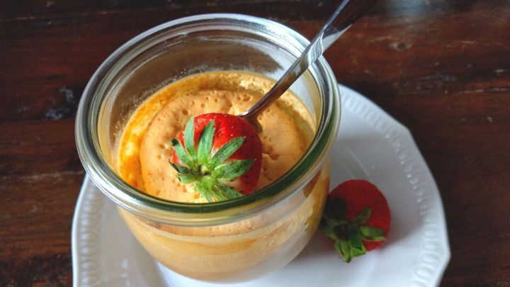 Ein Glas mit K&auml;sekuchen und einem L&ouml;ffel steht auf einem Teller, dekoriert mit einer frischen Erdbeere.