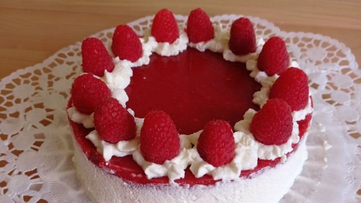 Eine kleine Himbeertorte steht auf einer Spitzenunterlage, dekoriert mit Sahne und frischen Himbeeren, vor einem hellen Hintergrund.