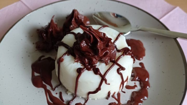Eine Panna Cotta mit Schokoso&szlig;e steht auf einem Teller, die Schokolade l&auml;uft dekorativ &uuml;ber die Creme.